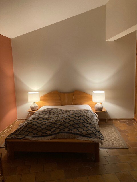 - Chambre cosy avec lit en bois, lampe chaleureuse, mur blanc et tapis beige sur sol carrelé.