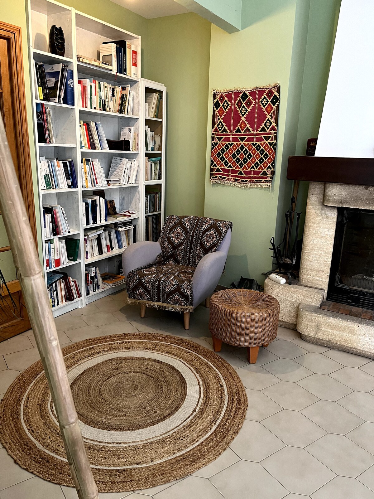 Coin lecture avec fauteuil, bibliothèque blanche, tapis tressé et cheminée en pierre.