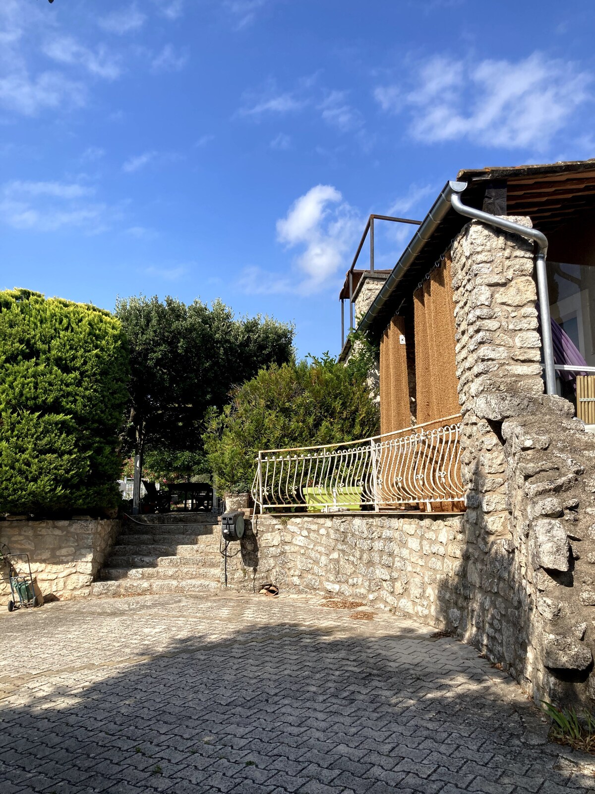 Cour en pierre avec escalier, balcon en fer forgé et végétation méditerranéenne sous ciel bleu.
