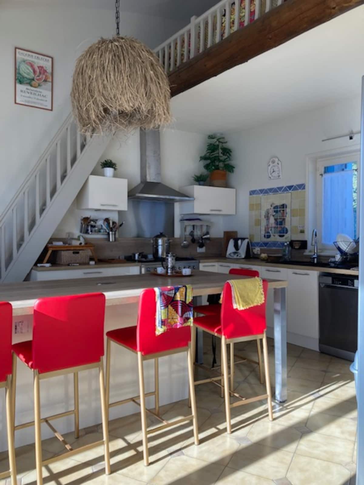 La cuisine de la Villa Naturasana allie modernité et charme rustique : îlot central en bois clair, chaises rouges, suspension en fibres naturelles, plantes en hauteur et détails décoratifs colorés. Un espace convivial et lumineux.