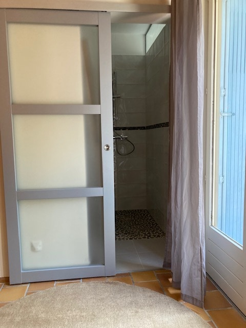 Douche moderne avec paroi vitrée, carrelage gris et sol en galets, éclairée par lumière naturelle.