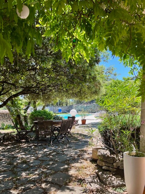 Jardin ombragé avec table en bois, chaises rustiques et piscine en arrière-plan.