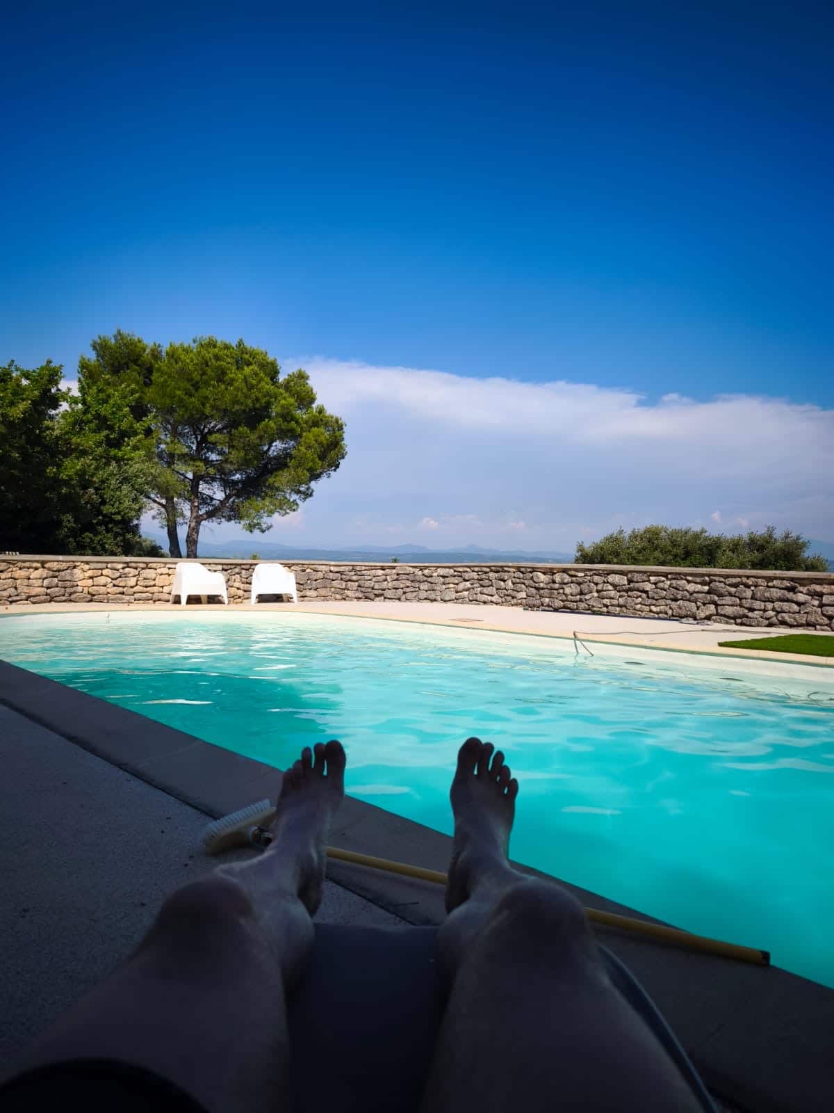 Personne détendue au bord de la piscine , avec vue sur transats, arbres et ciel bleu.