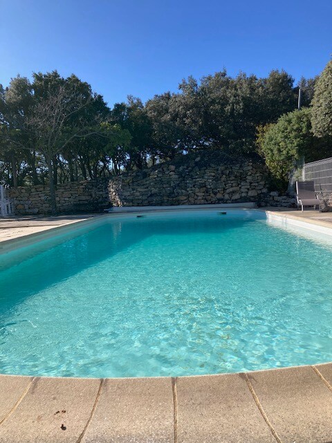 Piscine turquoise, entourée de pierres, arbres et transats, sous ciel bleu.