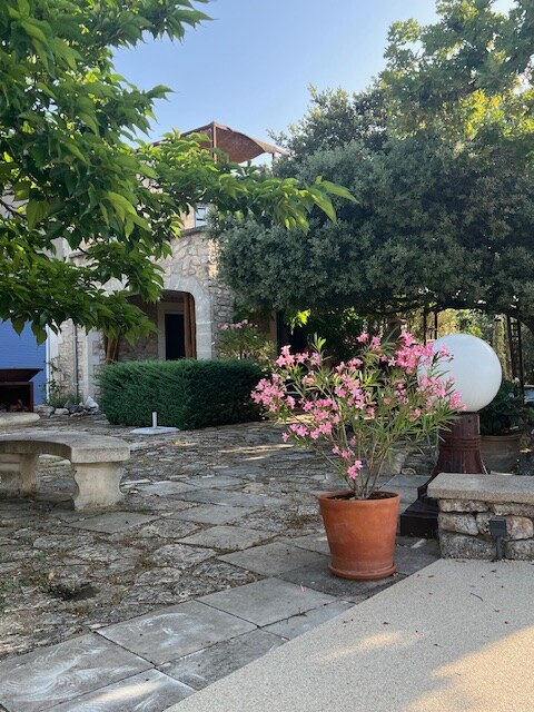 Pot de fleurs sur terrasse en pierre, banc et table basse devant maison en pierre ombragée.