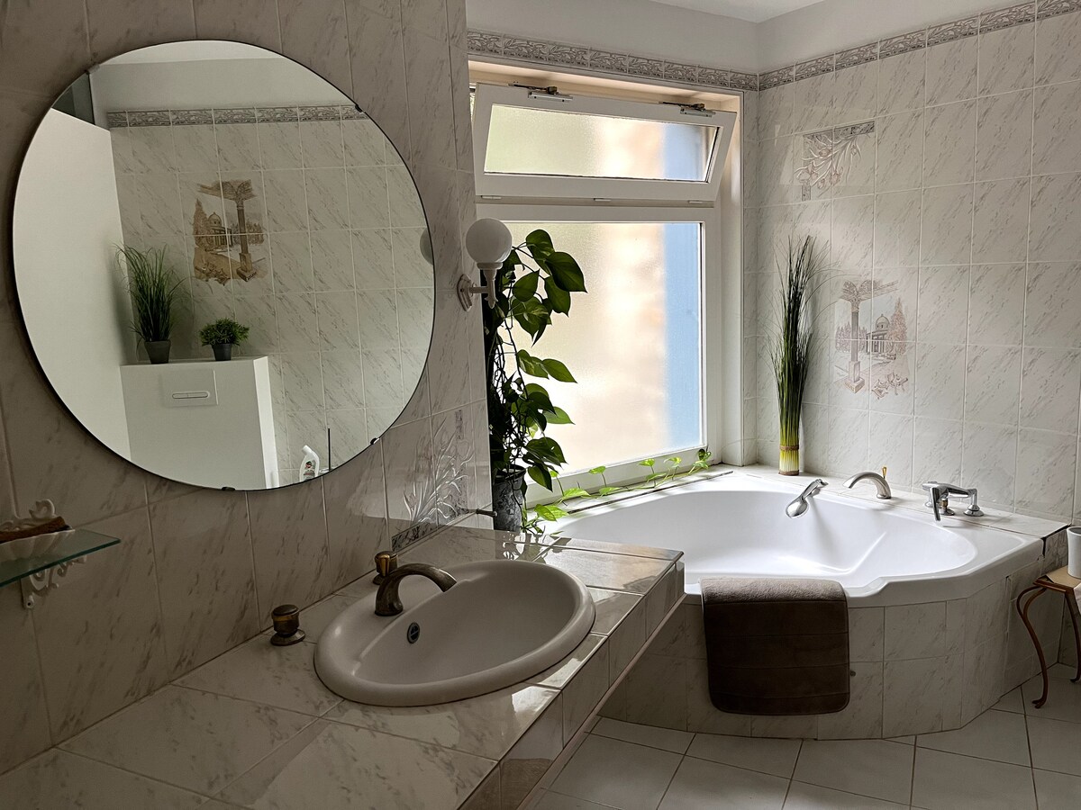 Salle de bain lumineuse avec baignoire d’angle, miroir rond et déco végétale sur fond marbré.
