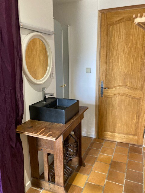 Salle d’eau avec vasque noire sur meuble ancien, miroir blanc et porte en bois naturel.