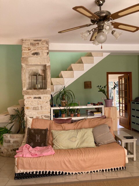 Salon confort  avec escalier blanc en pierre, canapé beige, plantes vertes et porte vitrée ouverte.