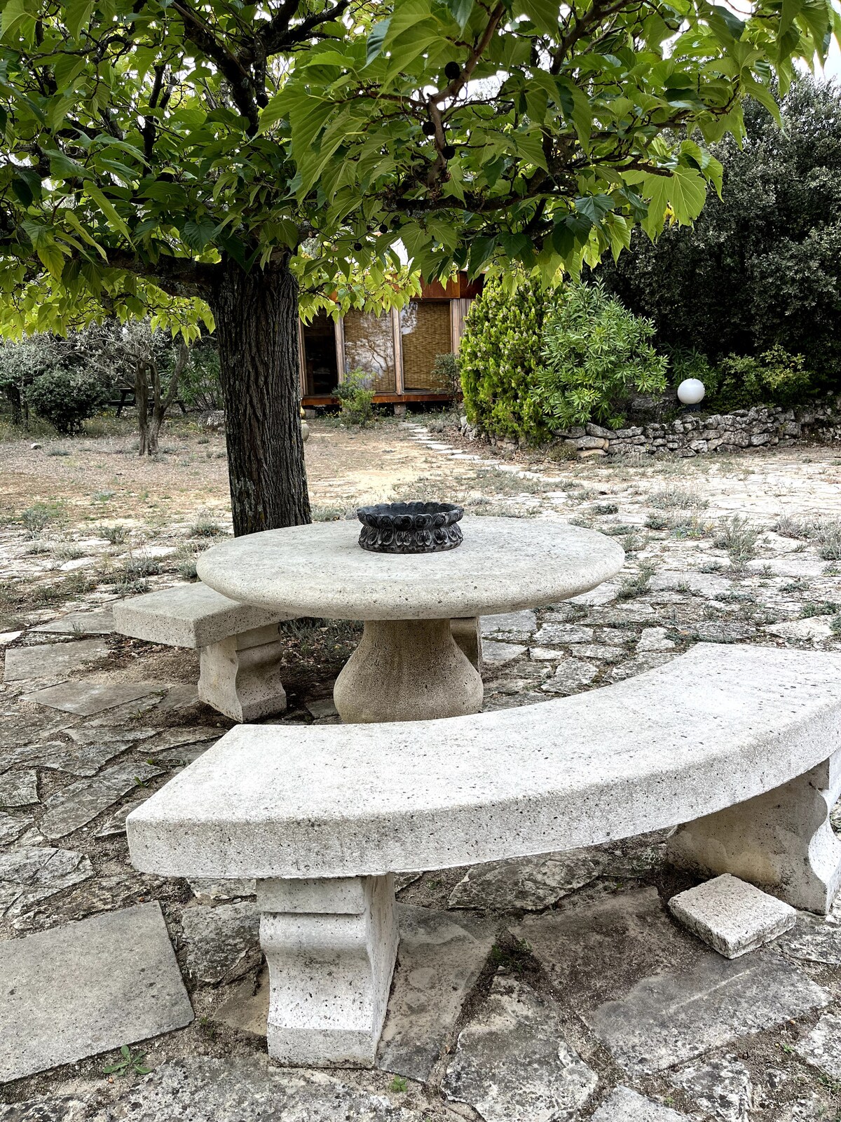 table ronde en pierre avec bancs courbes, sous arbre ombragé, près d’une yourte en bois.