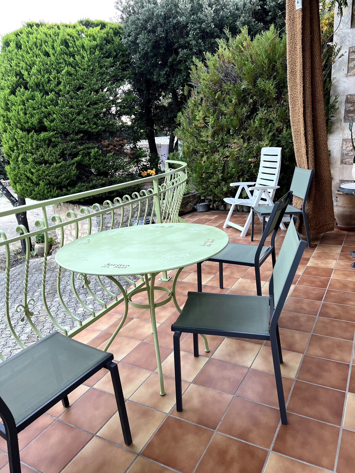 Terrasse carrelée avec table verte décorée, chaises dépareillées et végétation dense en arrière-plan.
