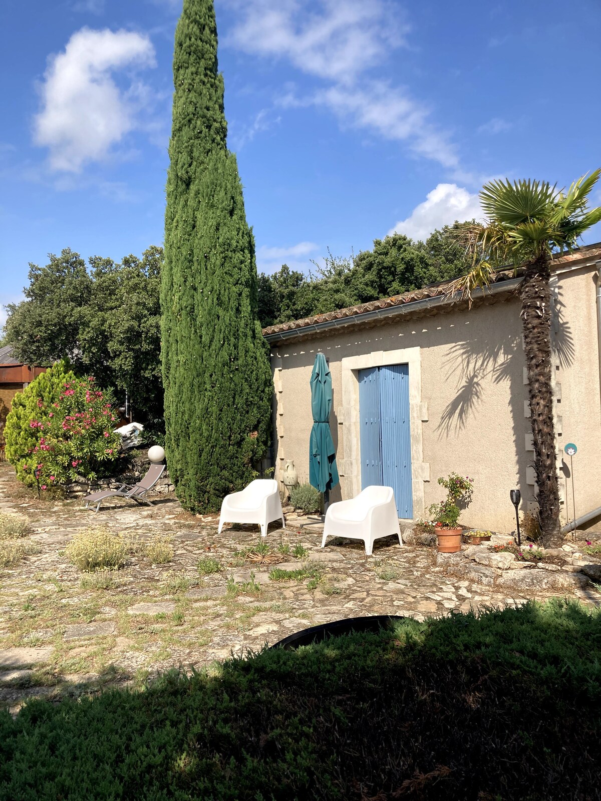 Terrasse en pierre avec chaises blanches, porte bleue et végétation méditerranéenne sous ciel dégagé.