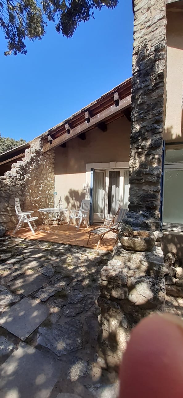 Terrasse en pierre avec table ronde et chaises blanches, devant maison en pierre aux volets clairs.