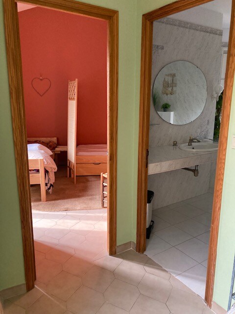 Vue intérieure sur chambre et salle de bain, murs verts, miroir rond et déco boisée.